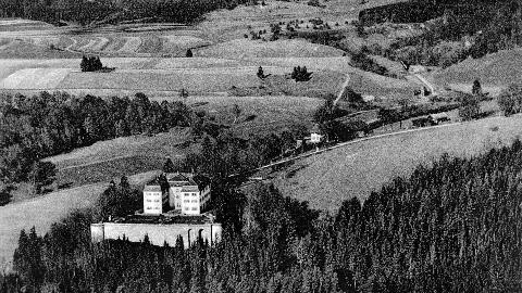 Historische Luftaufnahme des Schlosses und des gesamten Geländes in Grafeneck 1930. Im Vordergrund befindet sich das Schloss auf einer Anhöhe, umgeben von Wald. Im Hintergrund befindet sich das landwirtschaftliche Hauptgebäude und am rechten Bildrand das Gebäude, welches 1940 zur Gaskammer umgebaut wurde. Dahinter sind Felder und Bäume